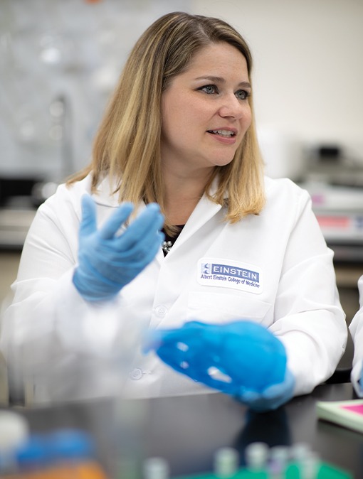 Woman with shoulder-length blond hair wearing white lab coat and blue gloves.