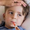Little boy with brown hair and brown eyes with fever thermometer in his mouth and adult's hand on his head.