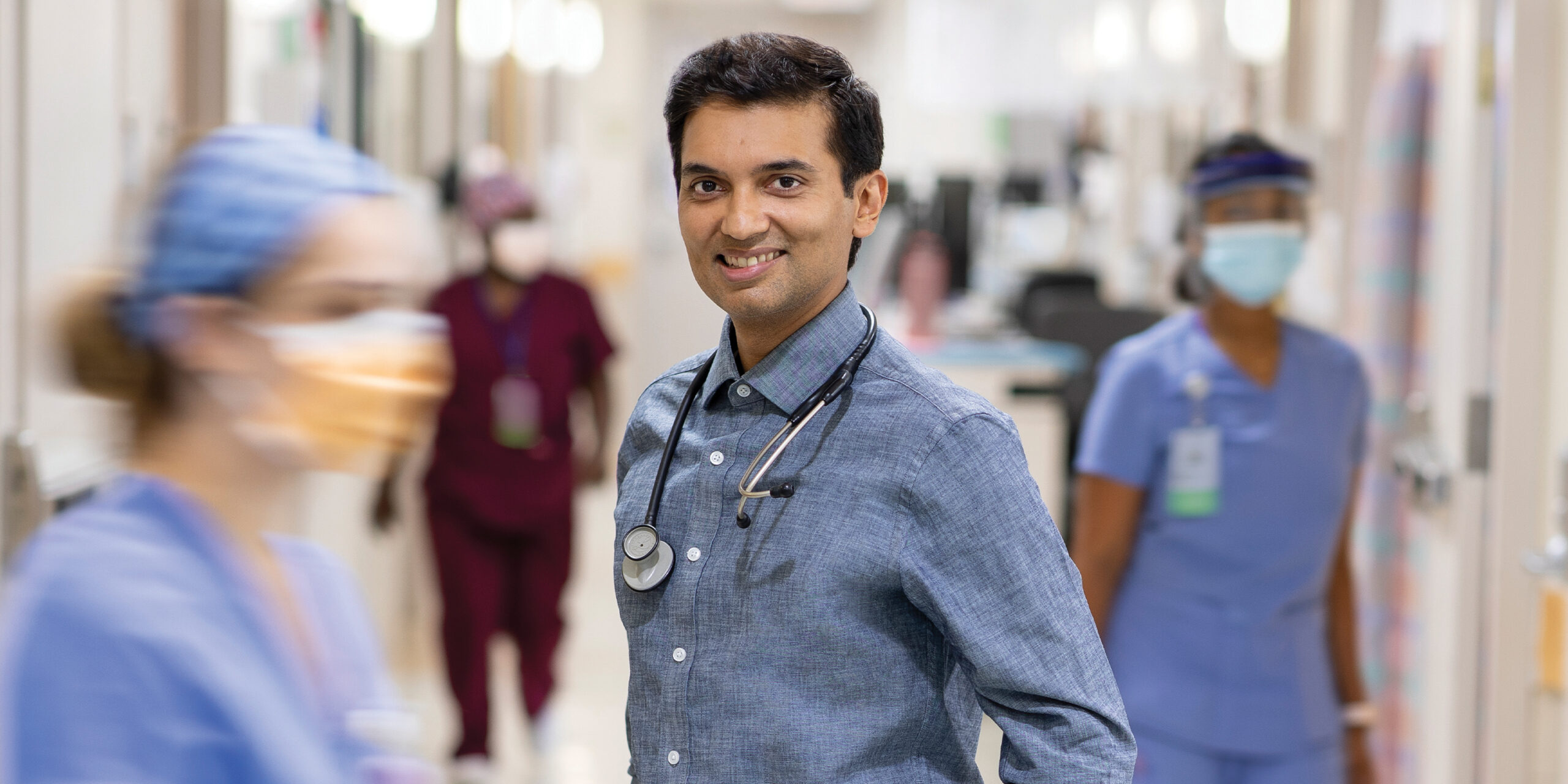 Man in blue shirt with stethoscope with hospital workers in background